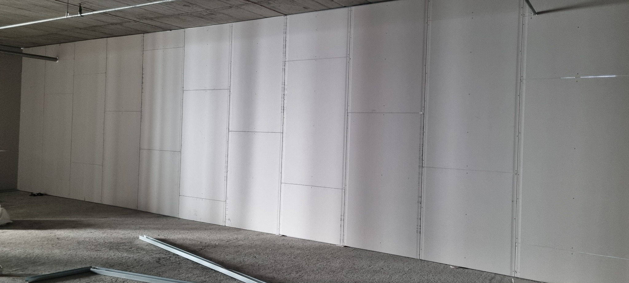 Ściana z płyt gipsowo-kartonowych w surowym wnętrzu, widoczne łączenia i metalowe profile na podłodze. Szare tło sufitu i podłogi podkreśla biel ścian.
