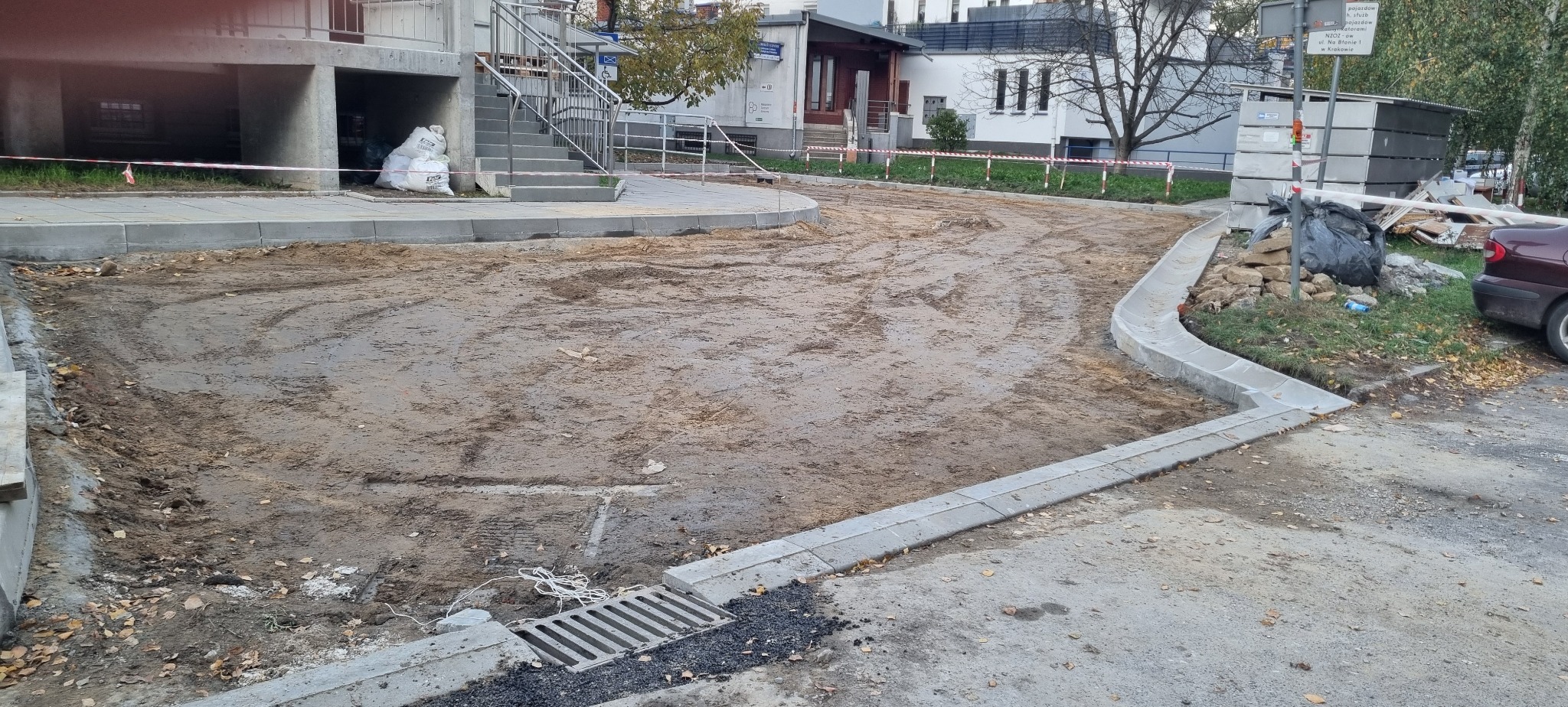 Teren po robotach ziemnych z nowymi krawężnikami i kratką ściekową. Widoczne ślady opon i przygotowany teren pod dalsze prace budowlane.