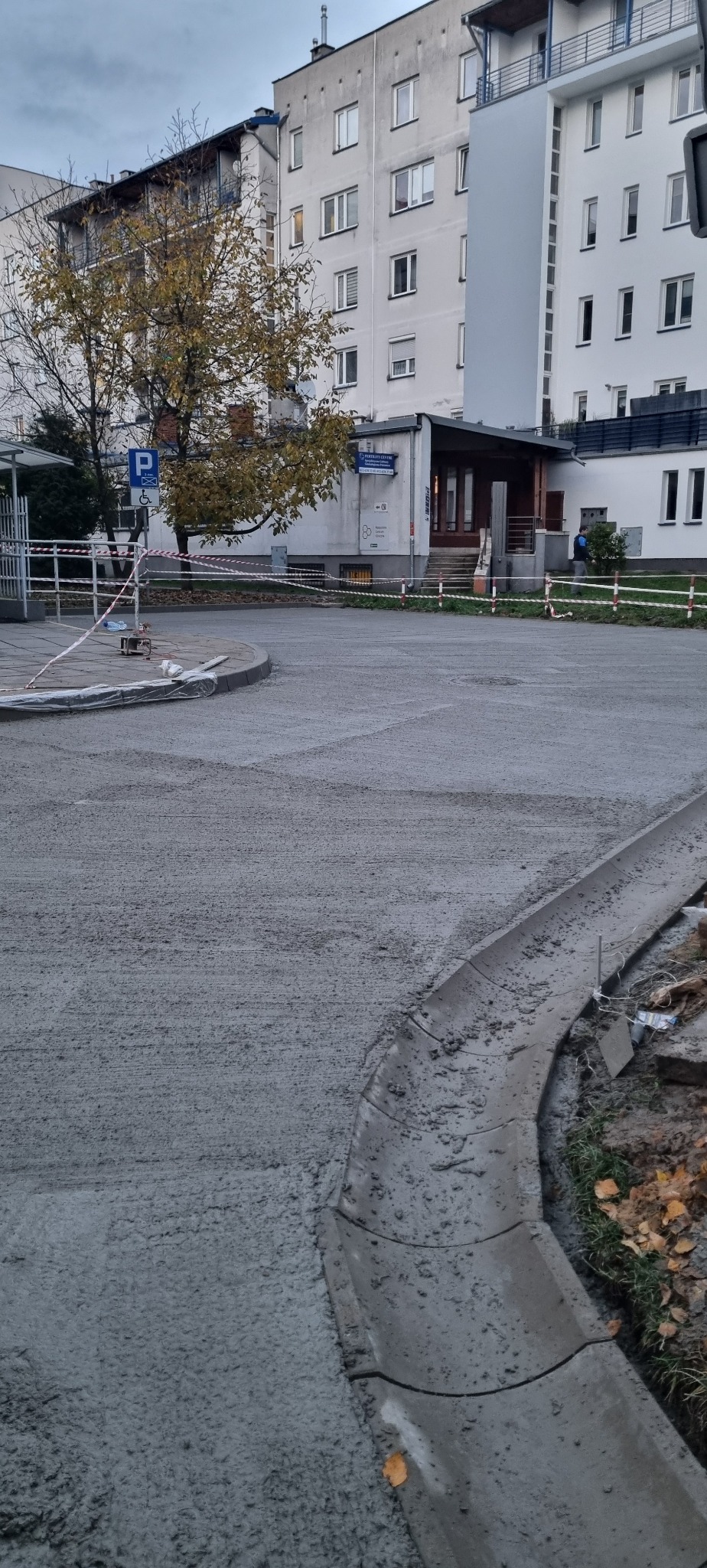 Świeżo wylana, szara wylewka na zewnątrz budynku, z widocznymi krawężnikami i taśmą ostrzegawczą. W tle budynki mieszkalne i drzewo z jesiennymi liśćmi.
