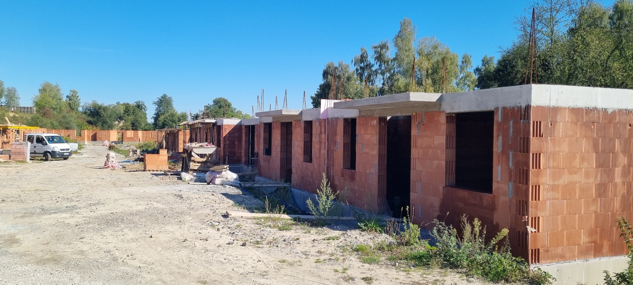 Wznoszenie szeregu budynków z czerwonej cegły, widoczne otwory okienne i betonowe nadproża. Na pierwszym planie gruz i roślinność, w tle zaparkowany biały bus.
