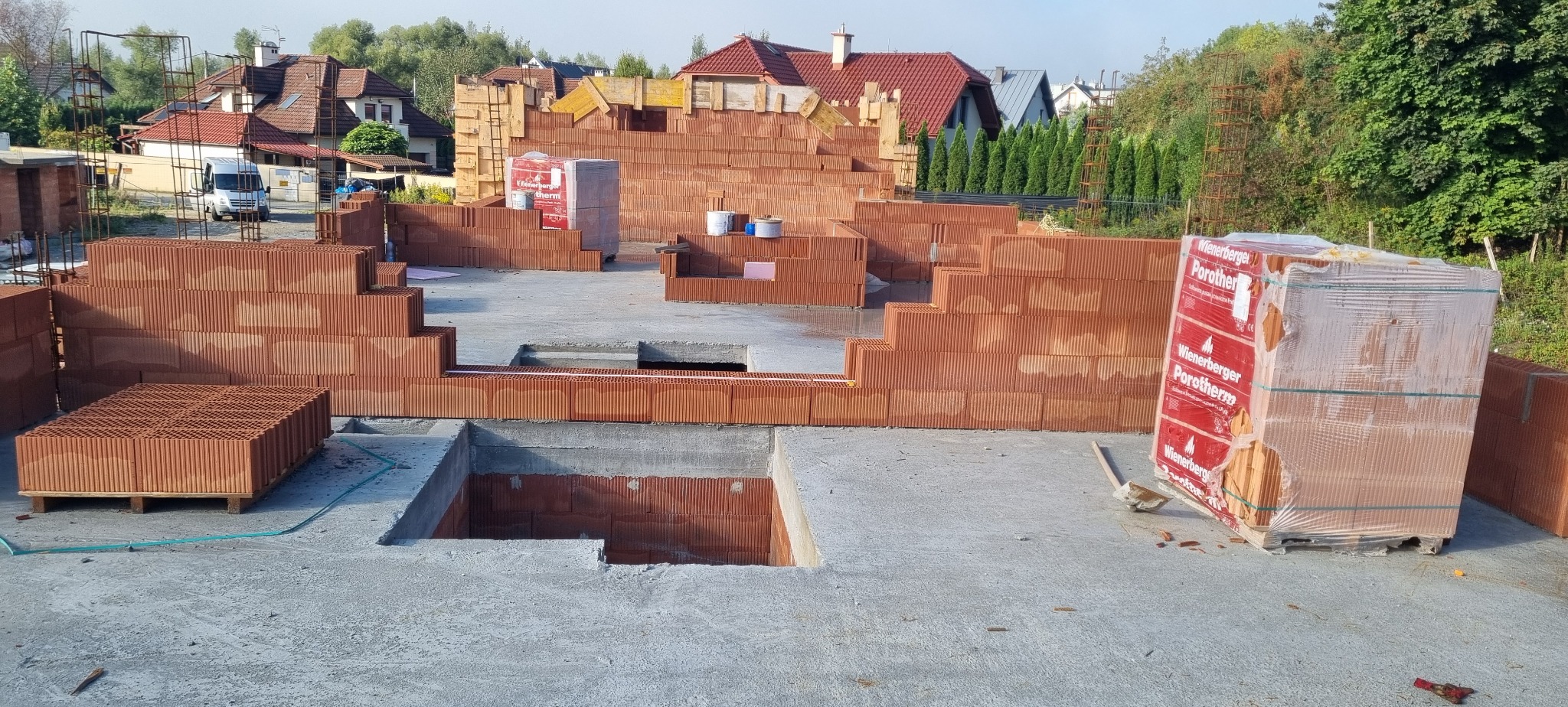 Budowa domu jednorodzinnego: fundamenty z wylewką i częściowo wymurowane ściany z czerwonej cegły Porotherm. Widoczne palety z cegłami i narzędzia budowlane.