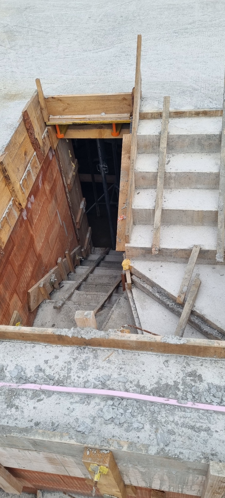 Betonowe schody w trakcie budowy z drewnianym szalunkiem, widok z góry. W tle widoczne rusztowania i elementy konstrukcyjne.