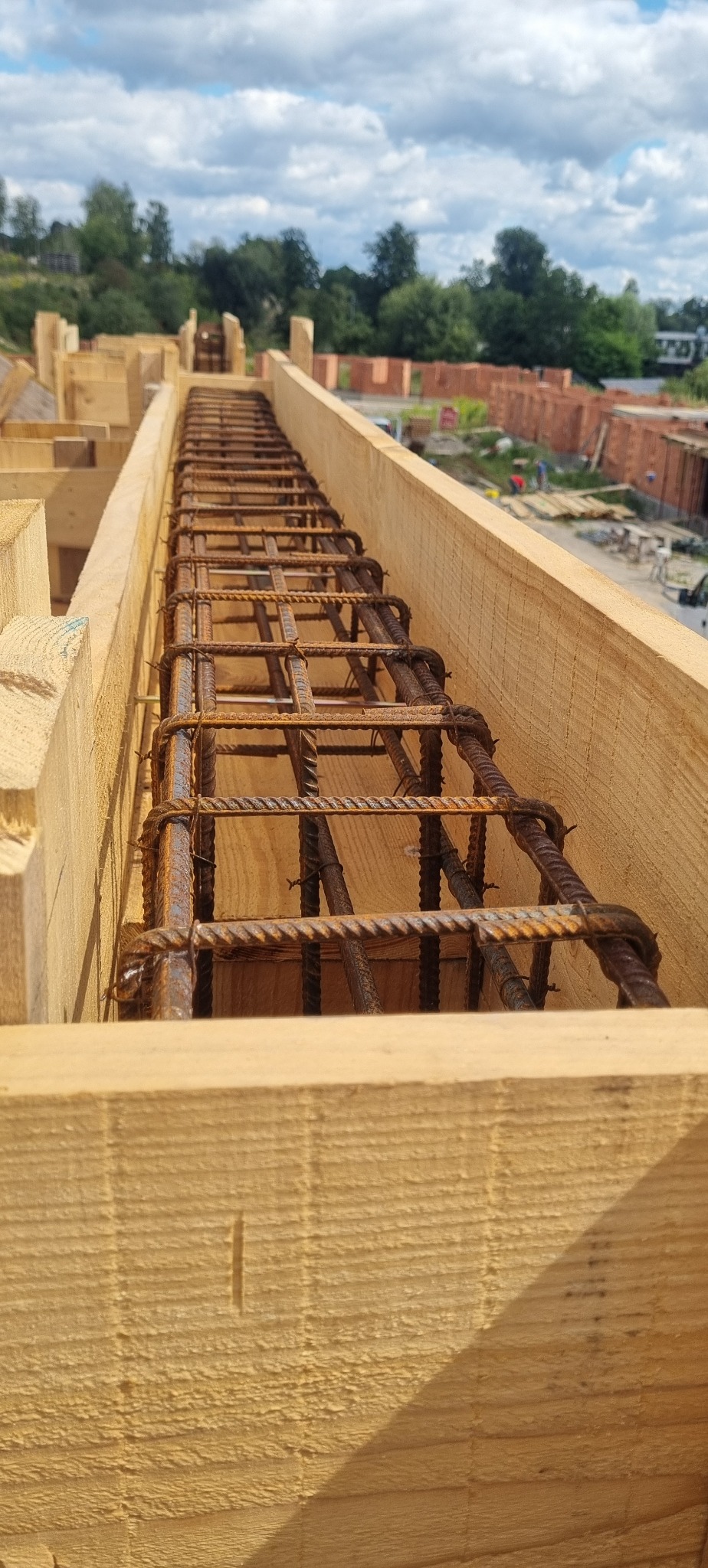 Zbrojenie w drewnianym szalunku, widok z góry. Na drugim planie ceglany budynek w budowie i zielone drzewa pod błękitnym niebem z chmurami.