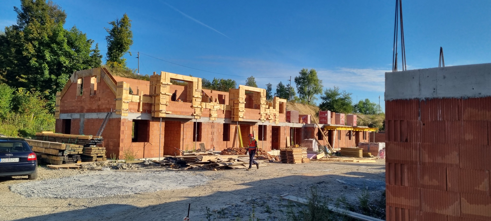 Budowa szeregówki z czerwonej cegły w Krakowie, widoczne drewniane szalunki okien i stropów, pracownik w kamizelce odblaskowej na placu budowy.