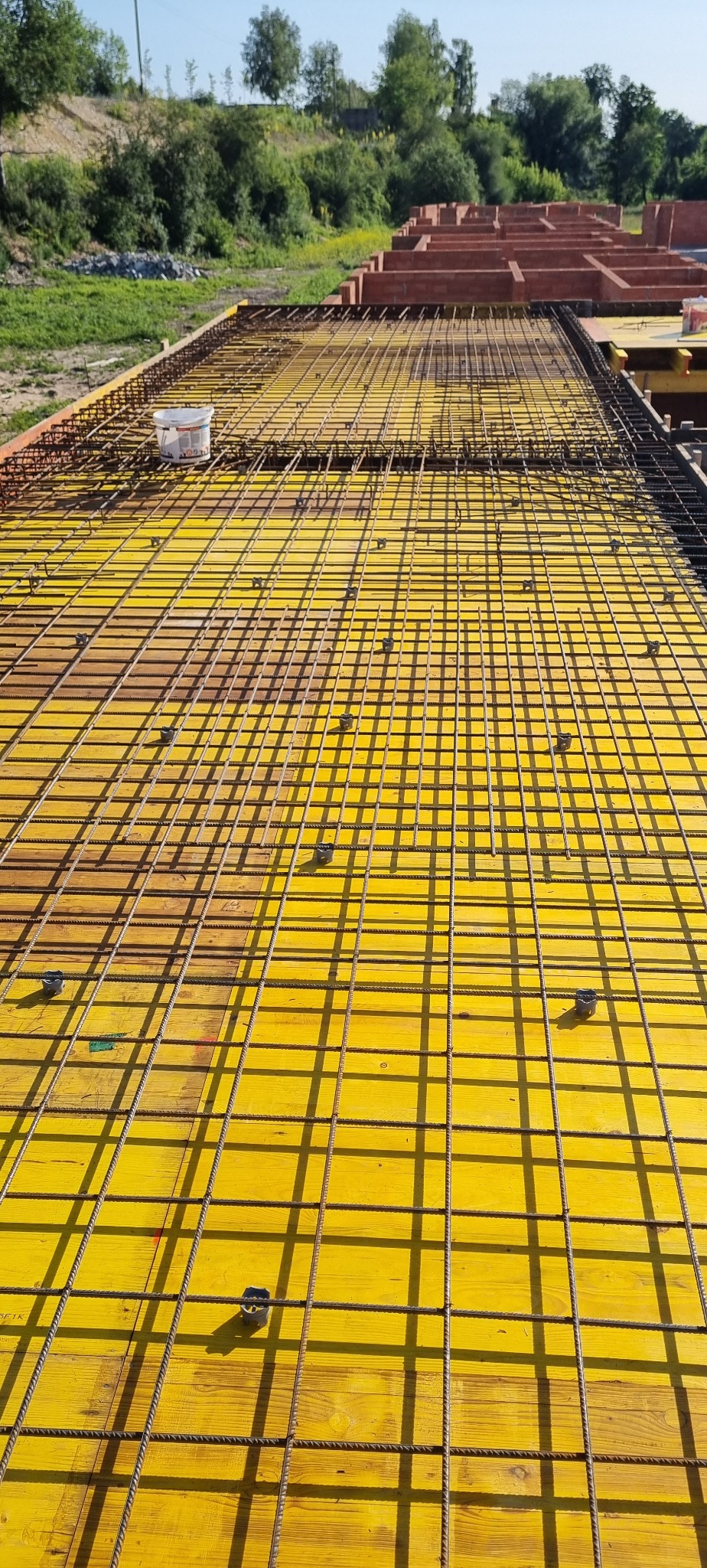 Żółty szalunek z siatką zbrojeniową i elementami mocującymi, widok z góry. W tle ceglane ściany i zieleń. Etap przygotowania do wylewki fundamentów.