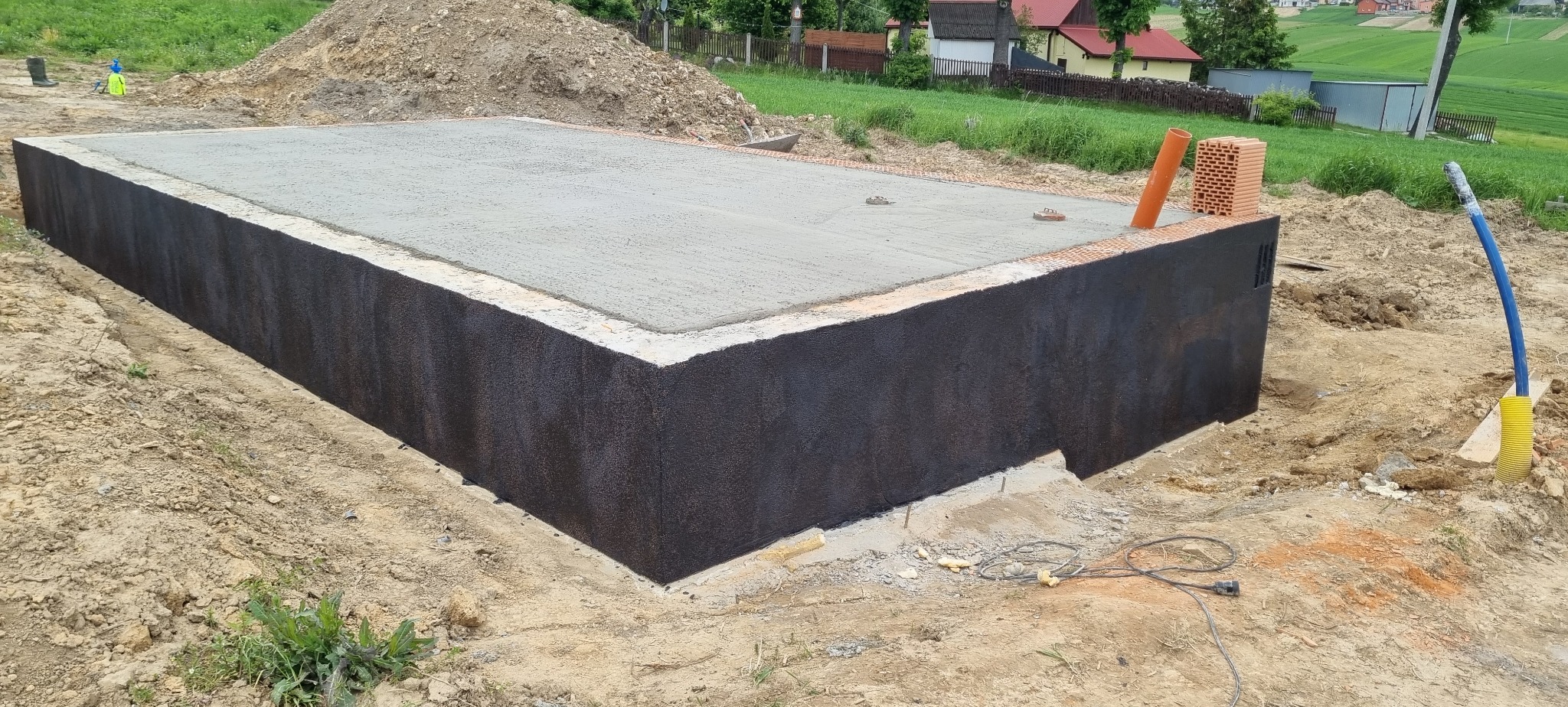 Zabezpieczona fundamentowa płyta betonowa z izolacją pionową, widoczne cegły i rury. W tle ziemia, trawa i zabudowania. Prace budowlane w toku.