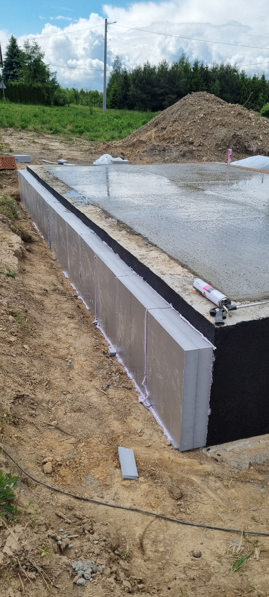 Fundamenty budynku w trakcie budowy: wylana płyta betonowa, ocieplenie pionowe styropianem, widoczna piana montażowa. W tle ziemia i zieleń.