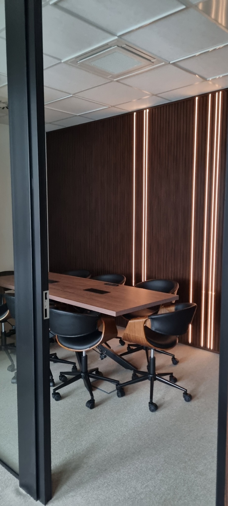 Nowoczesna sala konferencyjna z drewnianą ścianą z podświetleniem LED, czarnymi krzesłami i stołem. Widok przez otwarte drzwi, z widocznym fragmentem sufitu.