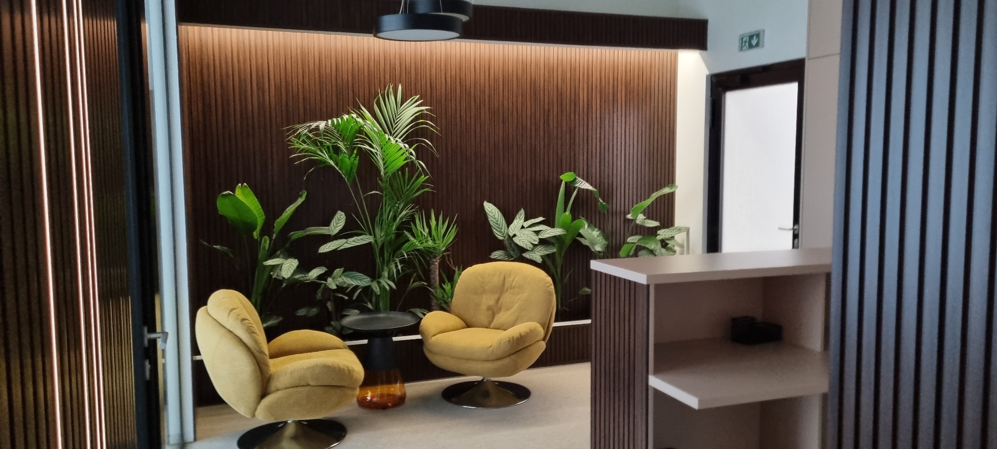 Nowoczesne wnętrze biurowe z dwoma fotelami, stolikiem i roślinami na tle drewnianej ściany z oświetleniem. Minimalistyczny design z elementami natury.