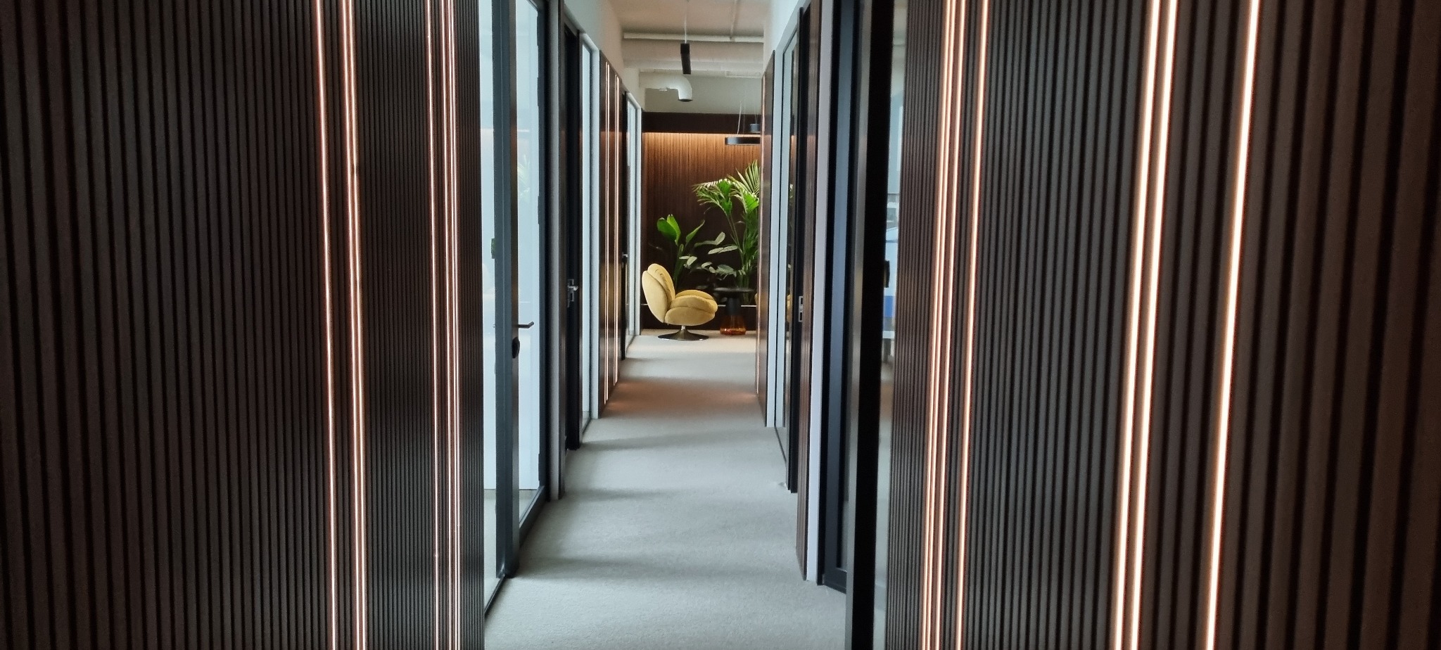 Korytarz biurowy z panelami ściennymi z podświetleniem LED, widok w głąb z żółtym fotelem i roślinami. Nowoczesne wykończenie wnętrza biura.