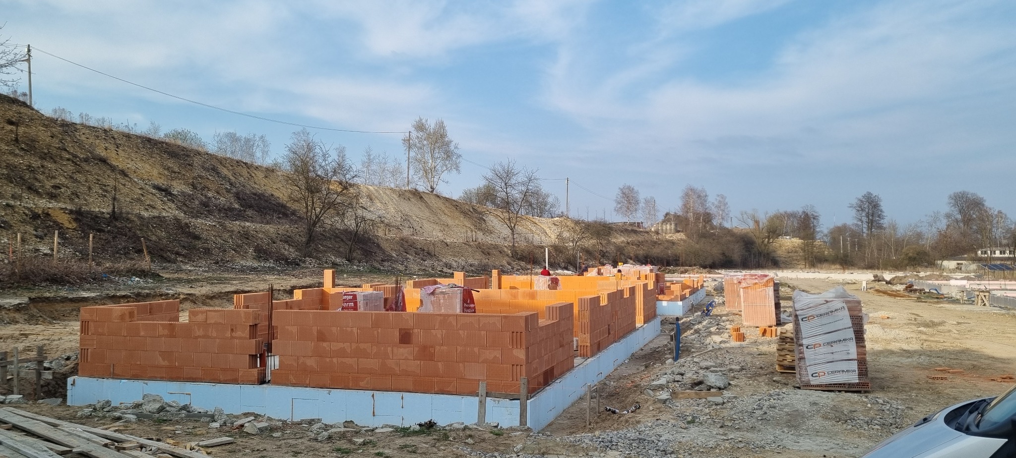 Wznoszenie ścian fundamentowych z czerwonej cegły na ocieplonym fundamencie. Widoczne palety z cegłami i teren budowy w tle.