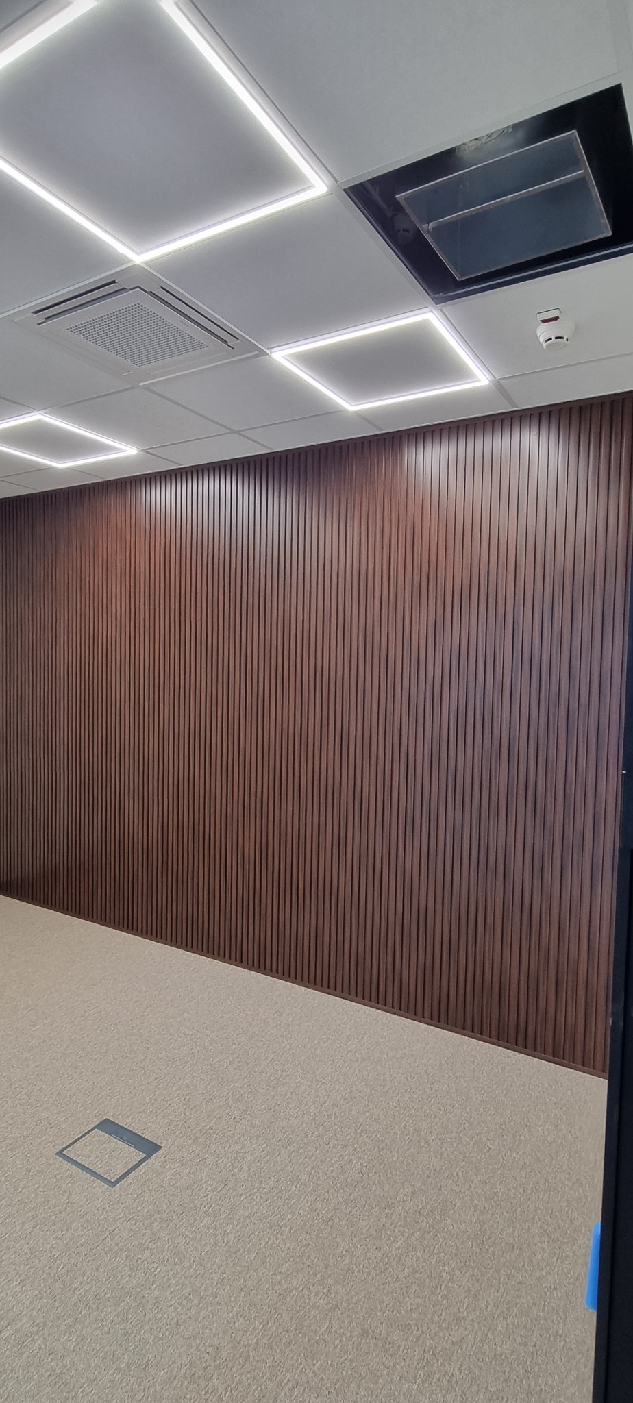 Nowoczesne wykończenie ściany w biurze z paneli drewnianych. Widoczne oświetlenie sufitowe LED i jasna wykładzina podłogowa. Minimalistyczny design wnętrza.