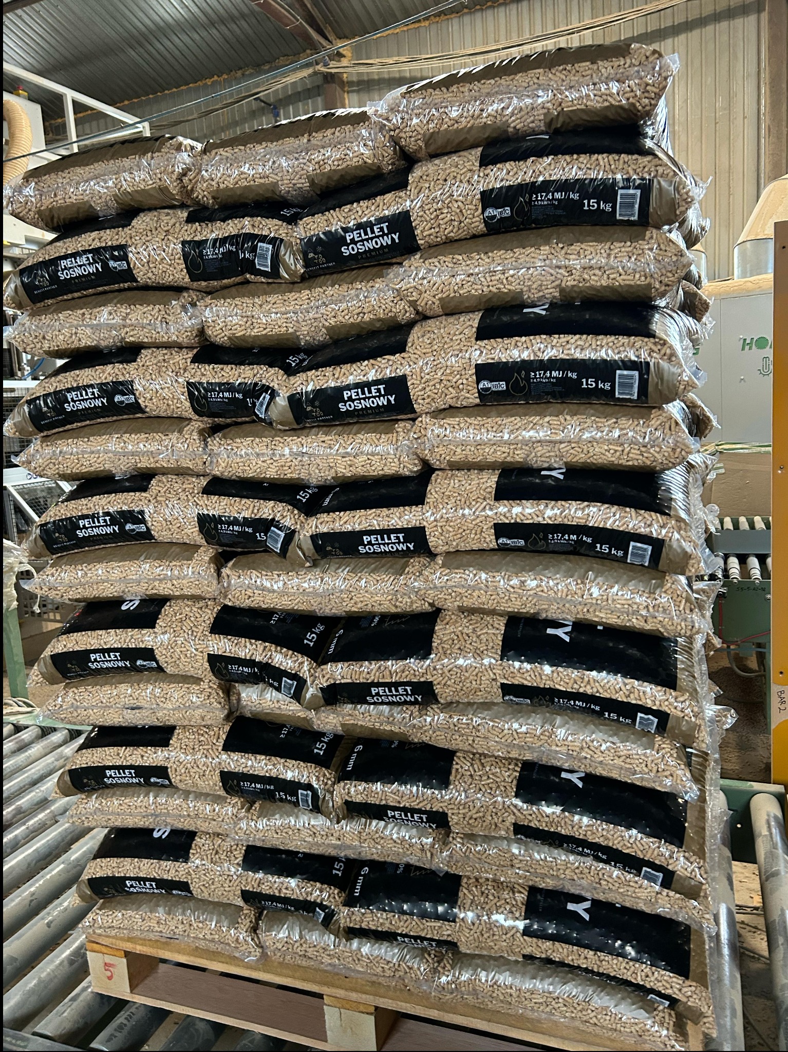 Stos worków z pelletem sosnowym, ułożonych na palecie w magazynie. Widoczne etykiety z informacjami o produkcie i wadze 15 kg. Tło stanowią elementy konstrukcji hali.