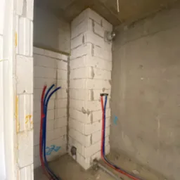Wnętrze w trakcie prac budowlanych: ściany z białych bloczków, rury instalacji wodnej w kolorze czerwonym i niebieskim, surowy beton na ścianie w tle.
