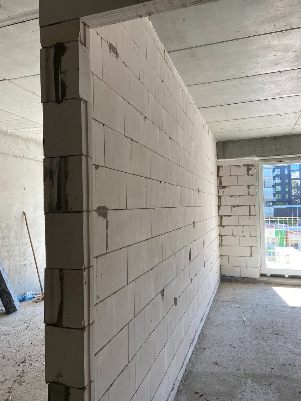 Świeżo postawiona ściana z białych bloczków z betonu komórkowego, widoczne spoiny i niedociągnięcia fugi, surowy stan deweloperski, okno w tle, betonowa podłoga.