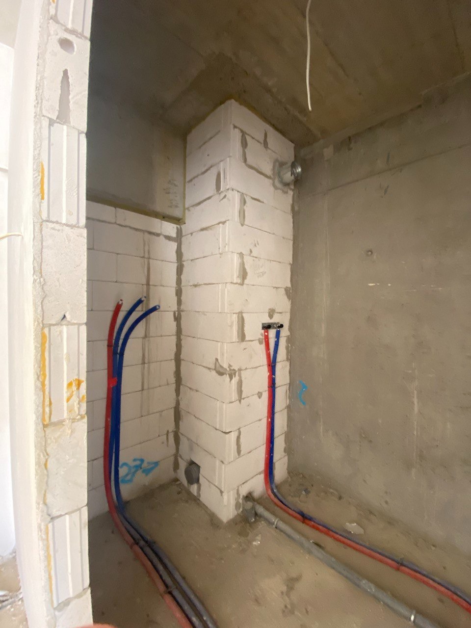 Wnętrze w trakcie prac budowlanych: ściany z białych bloczków, rury instalacji wodnej w kolorze czerwonym i niebieskim, surowy beton na ścianie w tle.