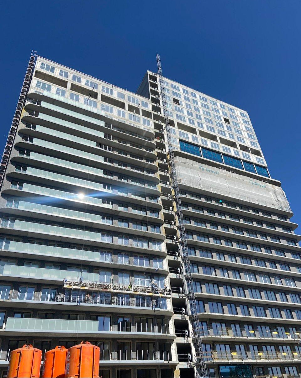 Wysoki budynek w trakcie budowy z widocznymi rusztowaniami, częściowo osłonięty siatką ochronną, z pomarańczowymi kontenerami na dole i intensywnym błękitem nieba w tle.