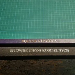 Dwa oprawione tomy książek, jeden fioletowy z napisem 'Rękopis z czyśćca', drugi czarny z napisem 'Śpiewnik pieśni kościelnych', leżące na zielonej macie introligatorskiej obok noży introligatorskich.