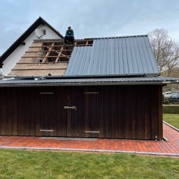 Robert ianek - Pracownik siedzi na dachu domu z częściowo zdjętym starym pokryciem i nową blachą trapezową, widoczne krokwie i łaty, na dachu antena satelitarna, w tle drzewo bez liści i pochmurne niebo.