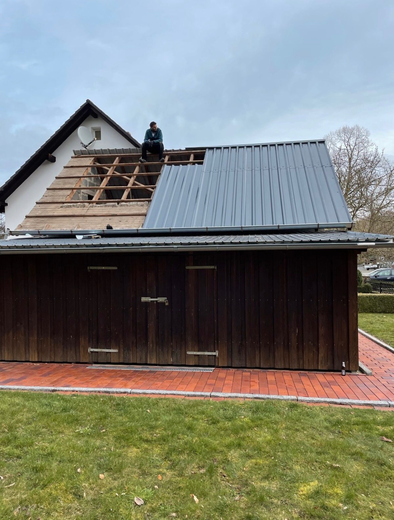 Pracownik siedzi na dachu domu z częściowo zdjętym starym pokryciem i nową blachą trapezową, widoczne krokwie i łaty, na dachu antena satelitarna, w tle drzewo bez liści i pochmurne niebo.