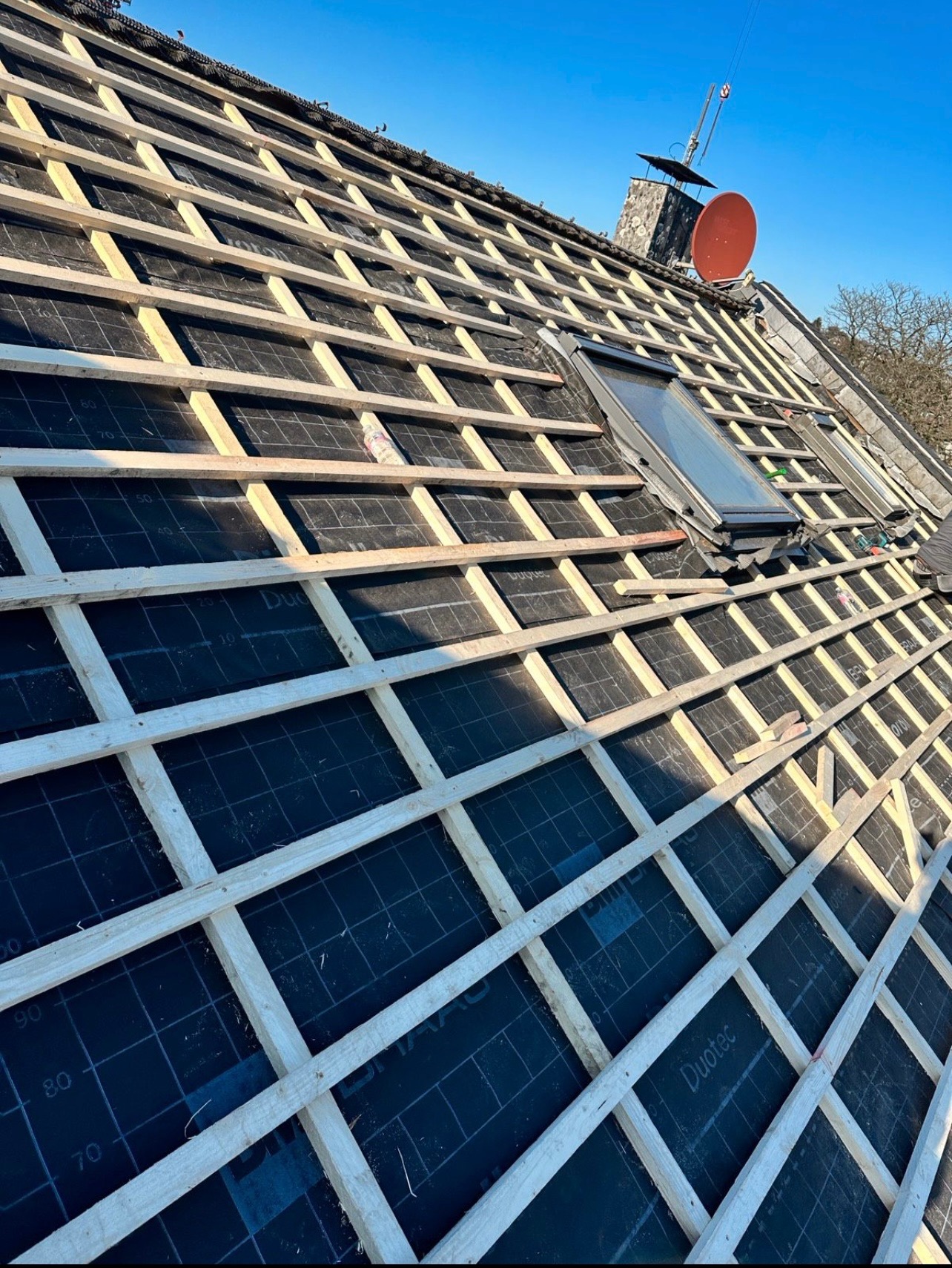 Konstrukcja drewniana łat na dachu pokrytym czarną membraną, widoczne okna dachowe i komin z anteną satelitarną na tle błękitnego nieba.