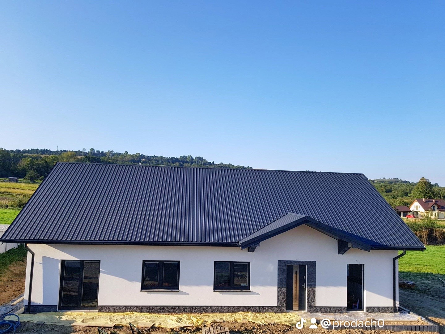 Nowoczesny dom jednorodzinny z ciemnym, metalowym dachem trapezowym na tle zielonych wzgórz i błękitnego nieba, widoczne czarne okna i obróbki blacharskie.