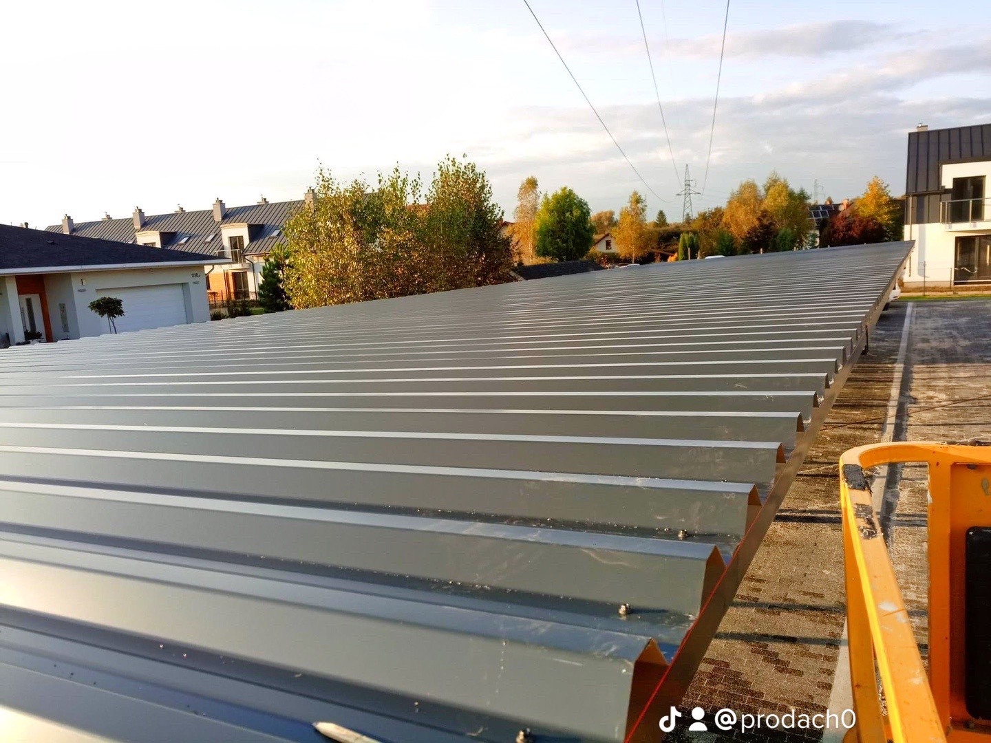 Widok z góry na szare, trapezowe blachy dachowe, częściowo zamontowane na konstrukcji, z widocznymi budynkami mieszkalnymi w tle i fragmentem podnośnika koszowego po prawej stronie.