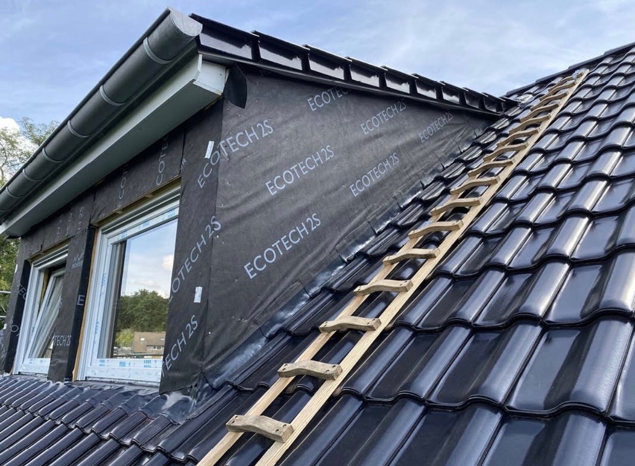 Częściowy widok dachu z czarną dachówką, zabezpieczonego membraną z nadrukiem 'ECOTECH 2S' przy oknie dachowym, z drewnianą drabiną dekarską opartą o połać dachu.
