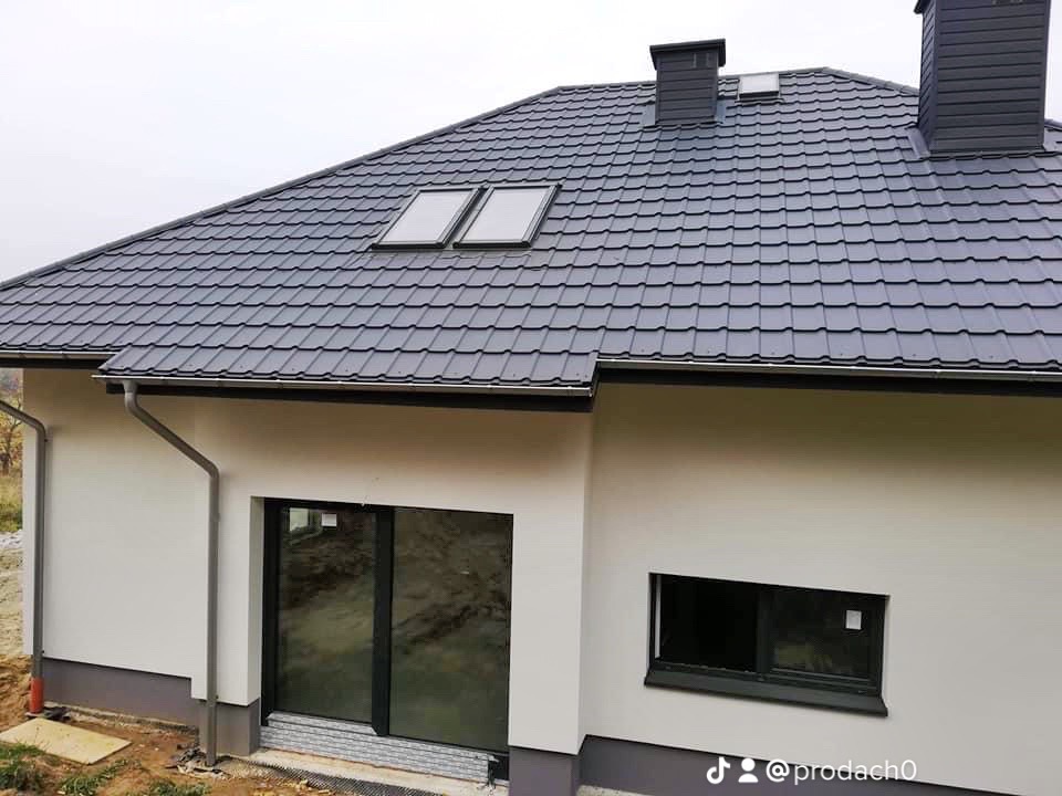 Nowy dom z grafitowym dachem z dachówki modułowej, dwoma oknami dachowymi i ciemnymi obróbkami blacharskimi, widok z zewnątrz w pochmurny dzień.