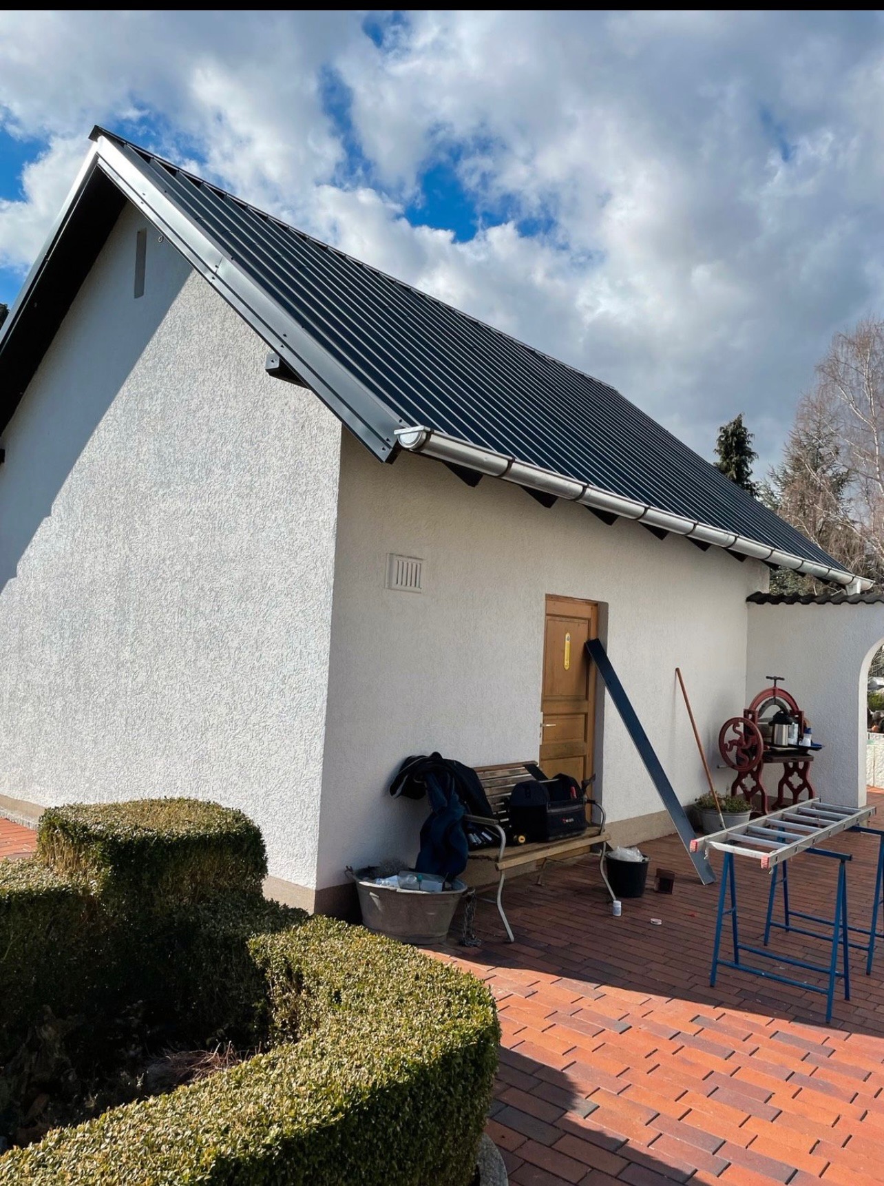 Nowy, czarny dach z blachy na białym budynku z widocznymi rynnami, narzędzia dekarskie i drabina na podwórku z kostki brukowej.