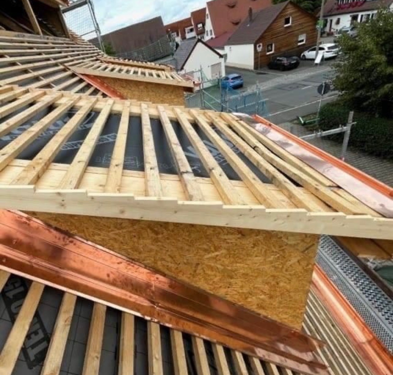Konstrukcja dachu w trakcie budowy: widoczne deski łat, kontrłaty, płyta OSB oraz elementy z blachy miedzianej, z widokiem na zabudowę mieszkalną w tle.