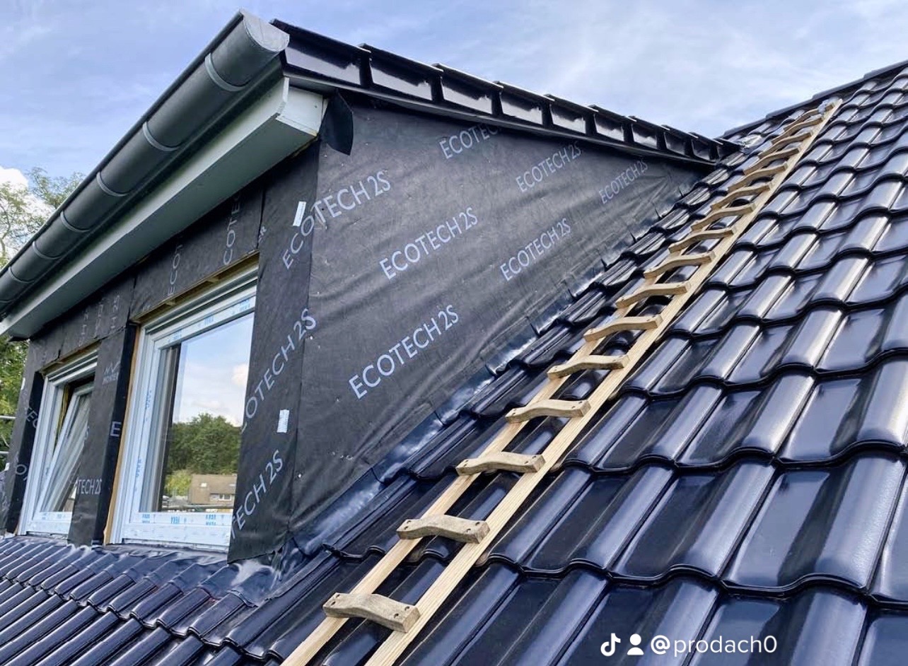 Częściowy widok dachu z czarnej dachówki ceramicznej, na którym leży drewniana drabina dekarska; widoczna folia dachowa z nadrukiem 'ECOTECH 2S' przy oknie dachowym i orynnowanie.