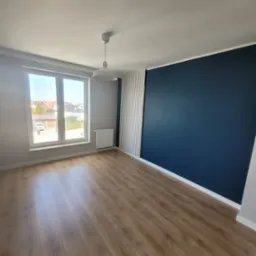 Pomalowane na granatowo wnętrze pokoju z panelami na podłodze, białą ścianą z grzejnikiem pod oknem i dekoracyjnymi białymi listwami pionowymi na ścianie obok granatowej ściany.