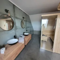 Nowoczesna łazienka na poddaszu z sauną, drewnianymi szafkami pod umywalki nablatowe, okrągłymi lustrami i szarymi płytkami imitującymi beton.