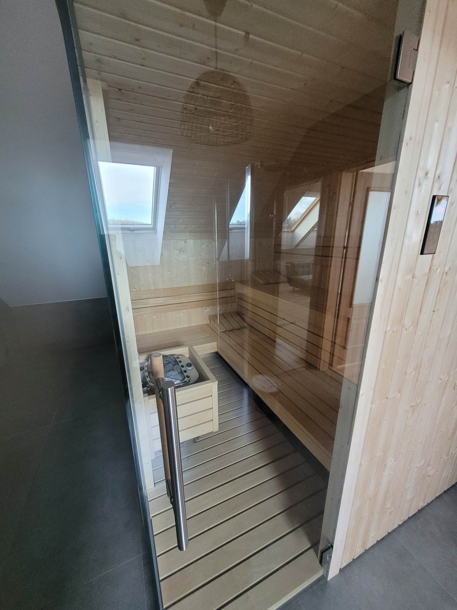 Nowoczesna sauna z drewnianym wnętrzem i szklanymi drzwiami, widok na poddasze z oknem dachowym, piec do sauny z kamieniami i metalowym uchwytem.