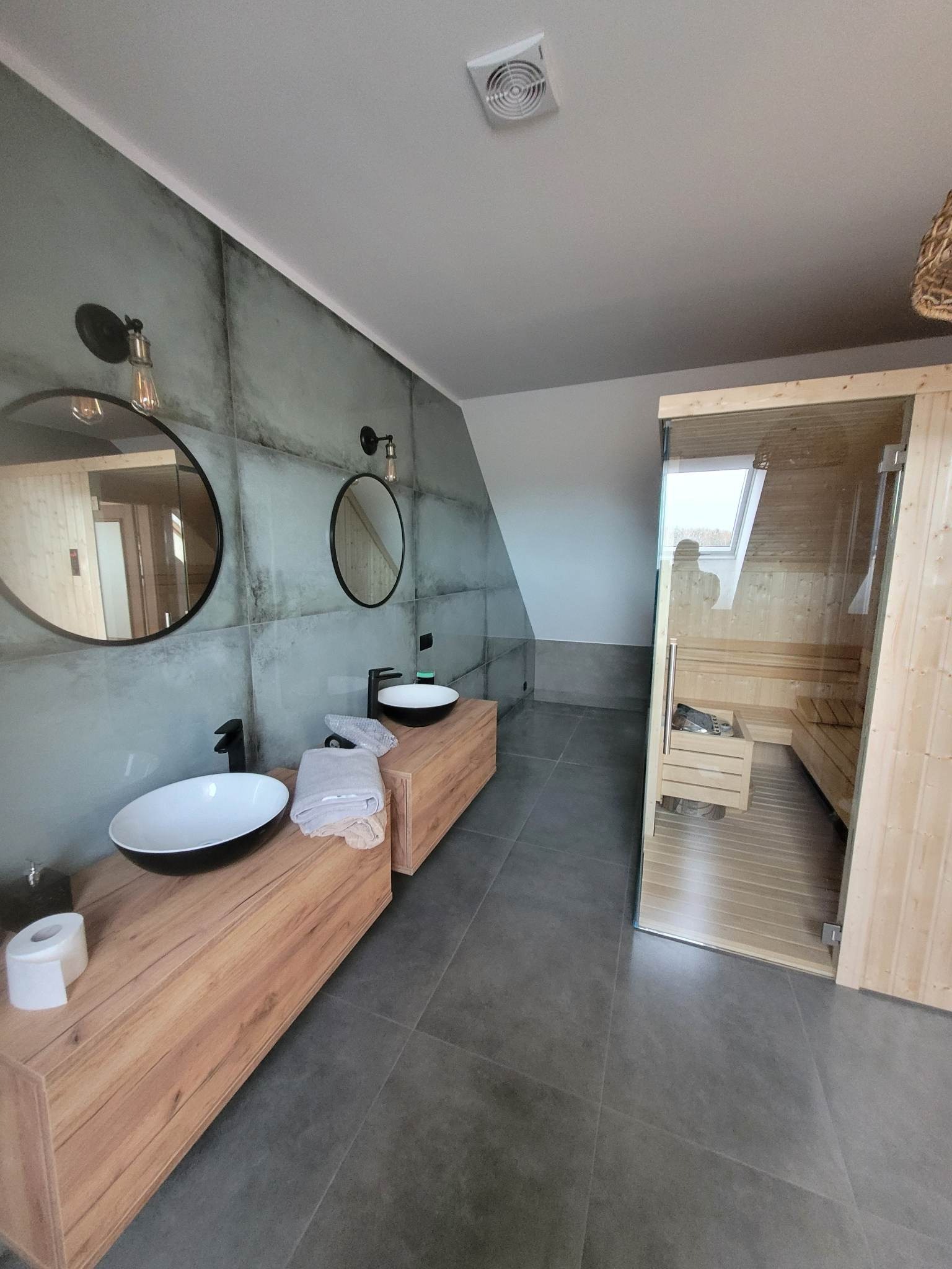 Nowoczesna łazienka na poddaszu z sauną, drewnianymi szafkami pod umywalki nablatowe, okrągłymi lustrami i szarymi płytkami imitującymi beton.