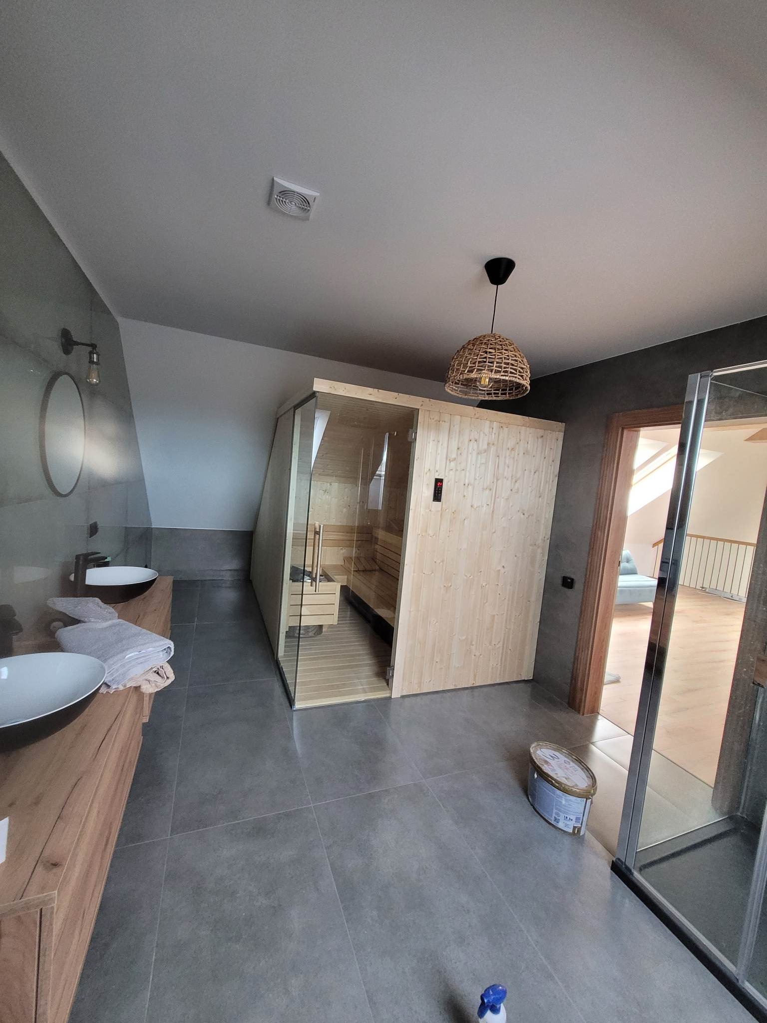 Nowoczesna łazienka z sauną, wykończona w szarościach, z drewnianymi akcentami, wiszącą lampą z abażurem i widocznym pojemnikiem z farbą na podłodze.