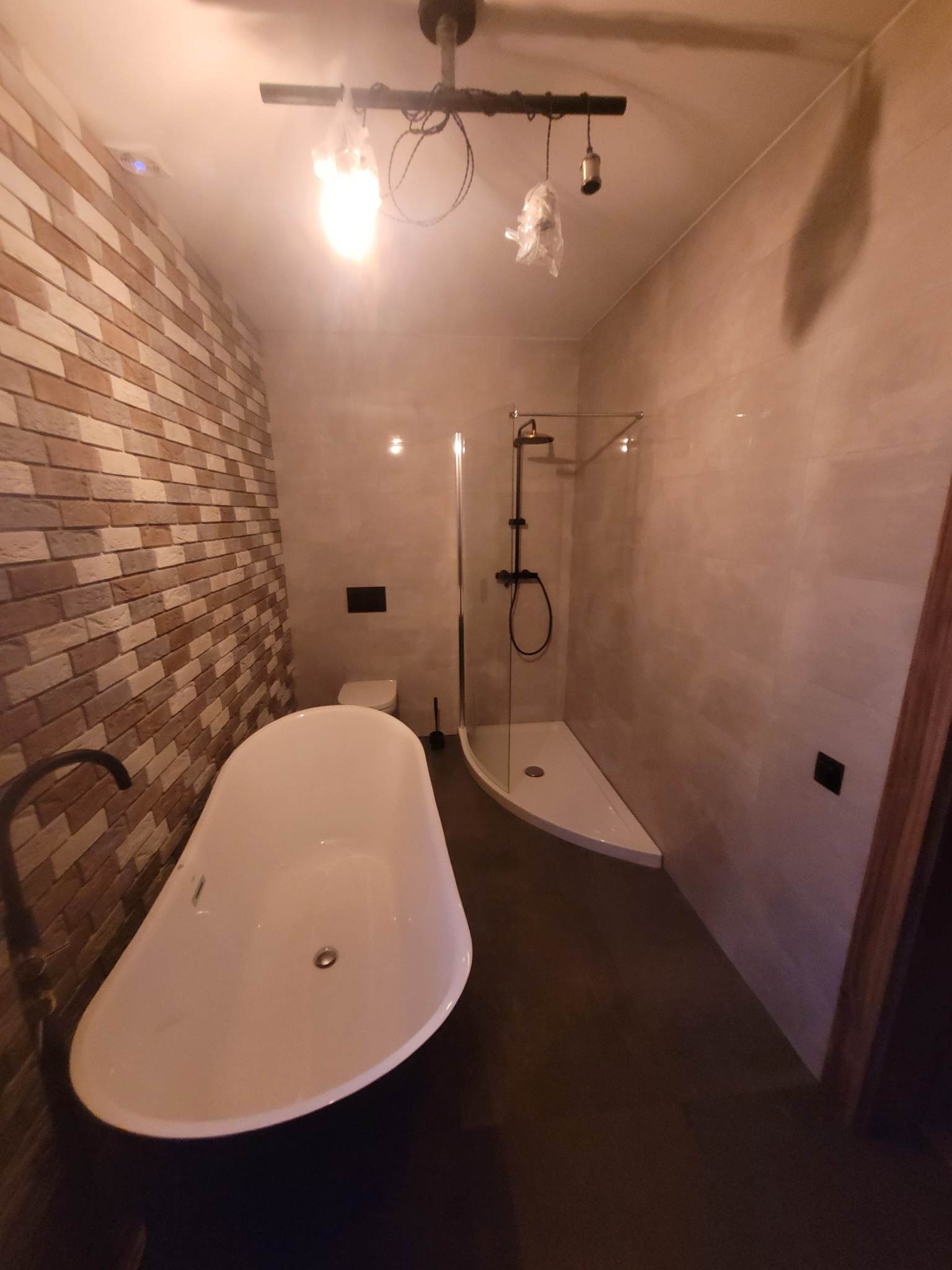 Nowoczesna łazienka z wolnostojącą wanną na ciemnej podłodze, ściana z cegieł, kabina prysznicowa ze szklaną ścianką i nietypowe oświetlenie sufitowe.