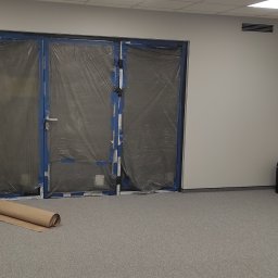 UPGRADE IVAN BOICHUK - Przygotowane pomieszczenie do malowania: zabezpieczone folią drzwi, rozłożona rolka papieru malarskiego, przedłużacz i spryskiwacz na podłodze z wykładziną dywanową.