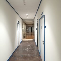 UPGRADE IVAN BOICHUK - Korytarz biurowy przygotowany do malowania: ściany i drzwi oklejone taśmą malarską, podłoga zabezpieczona folią ochronną, drabina w tle.