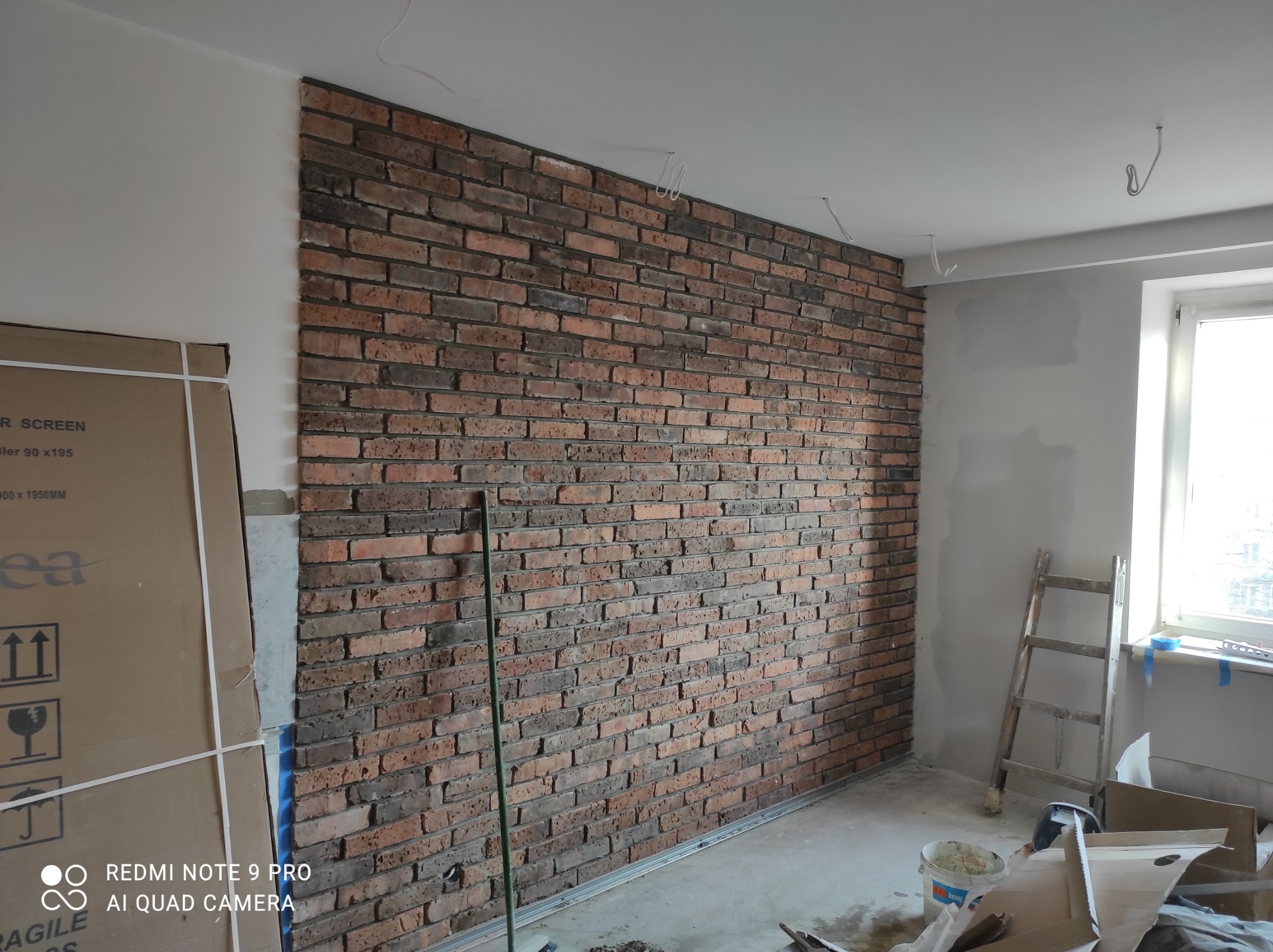 Ściana wyłożona cegłą klinkierową w trakcie prac wykończeniowych, widoczna drabina, wiadro z zaprawą i kartony w pomieszczeniu.