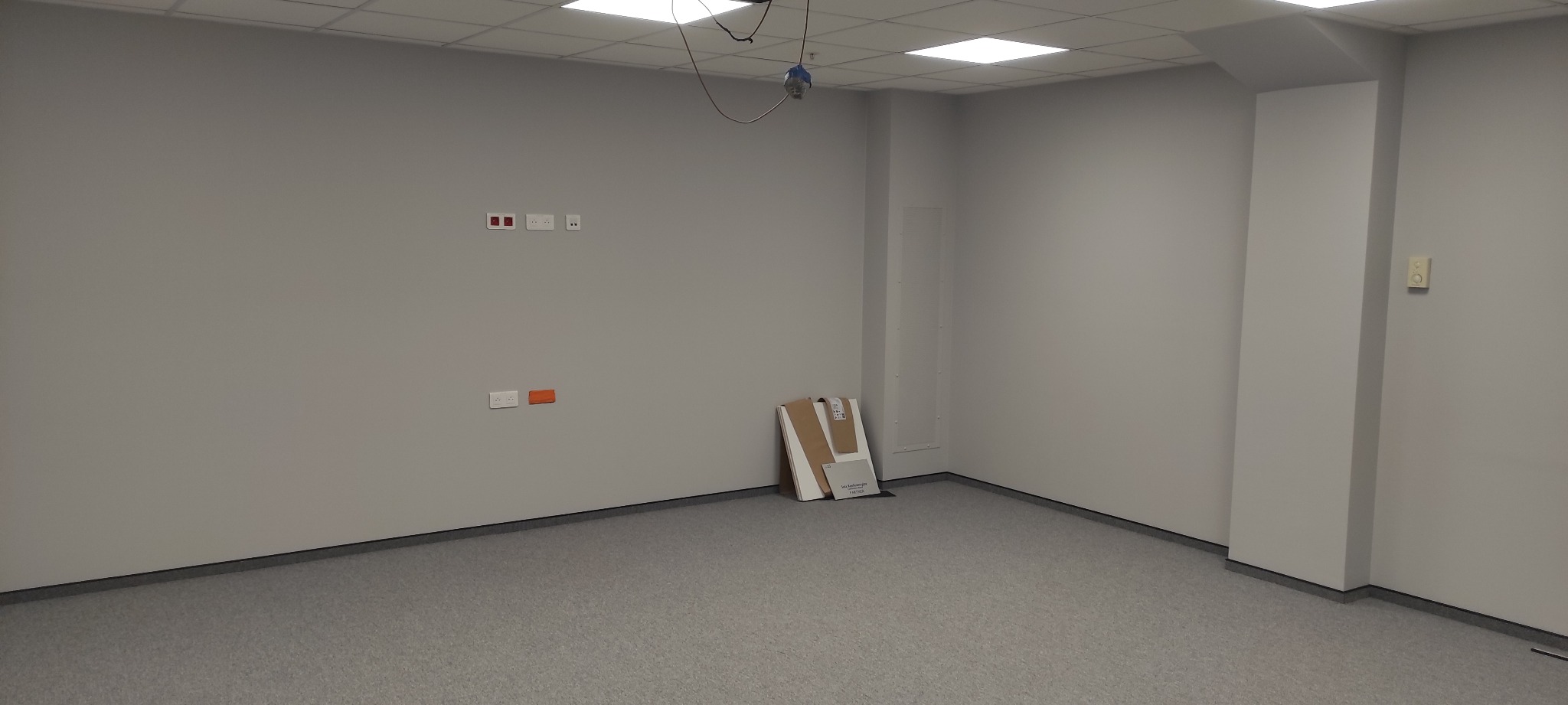 Pusty, świeżo pomalowany pokój biurowy z szarą wykładziną i gładkimi, szarymi ścianami, widoczne gniazdka elektryczne, wyłącznik i próbki materiałów przy ścianie.