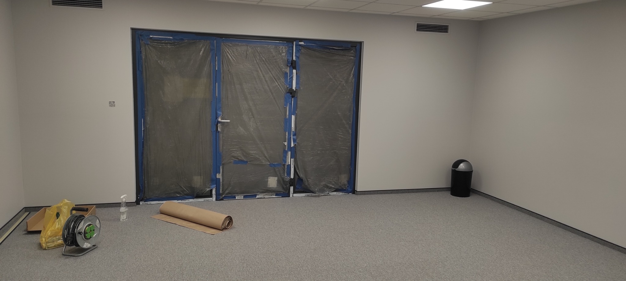 Przygotowane pomieszczenie do malowania: zabezpieczone folią drzwi, rozłożona rolka papieru malarskiego, przedłużacz i spryskiwacz na podłodze z wykładziną dywanową.