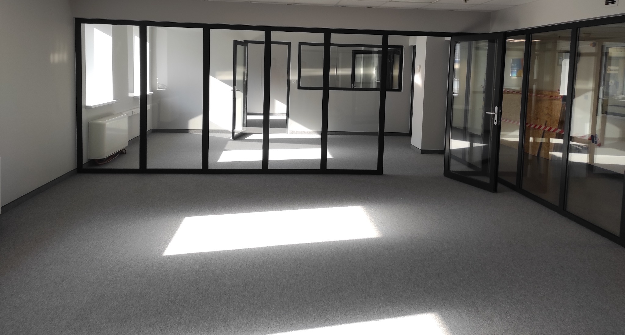Jasne, puste biuro z szarą wykładziną, dzielone szklanymi ścianami z czarnymi ramami, wpadające promienie słoneczne na podłogę.