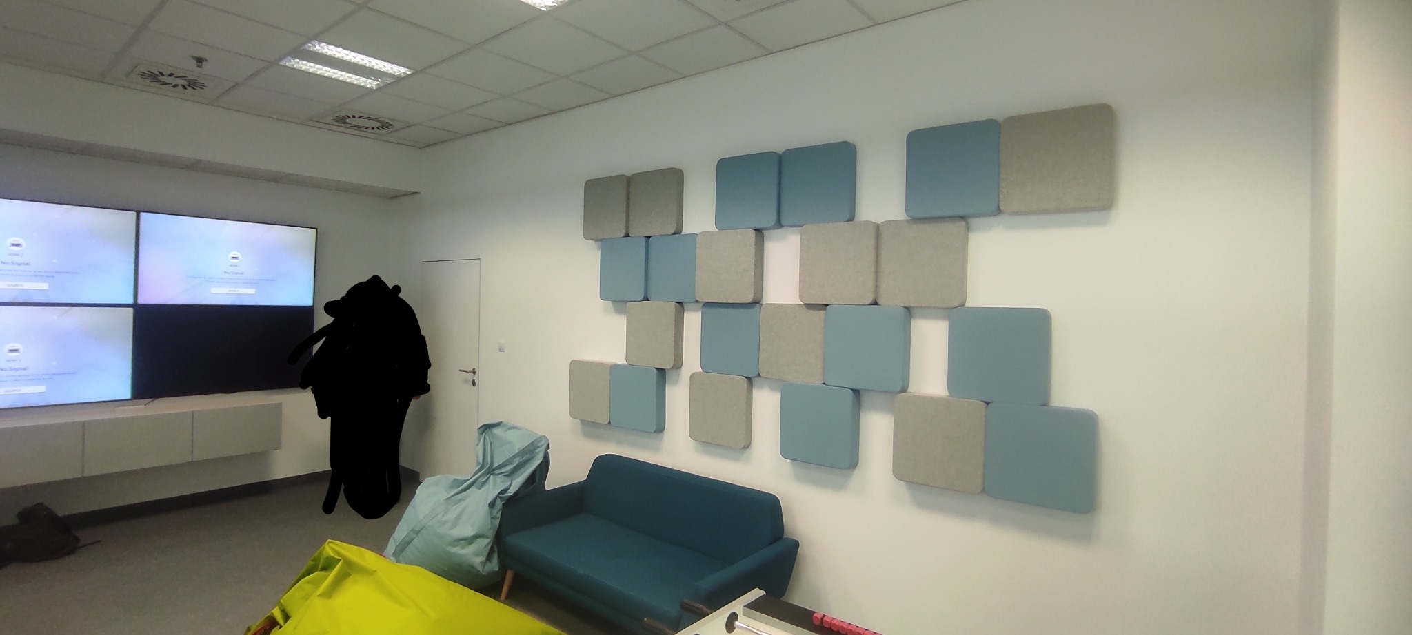 Ściana biurowa w Krakowie z dekoracyjnymi panelami akustycznymi w odcieniach szarości i błękitu, uzupełniona o niebieską sofę i cztery ekrany wyświetlające komunikat 'No signal'.