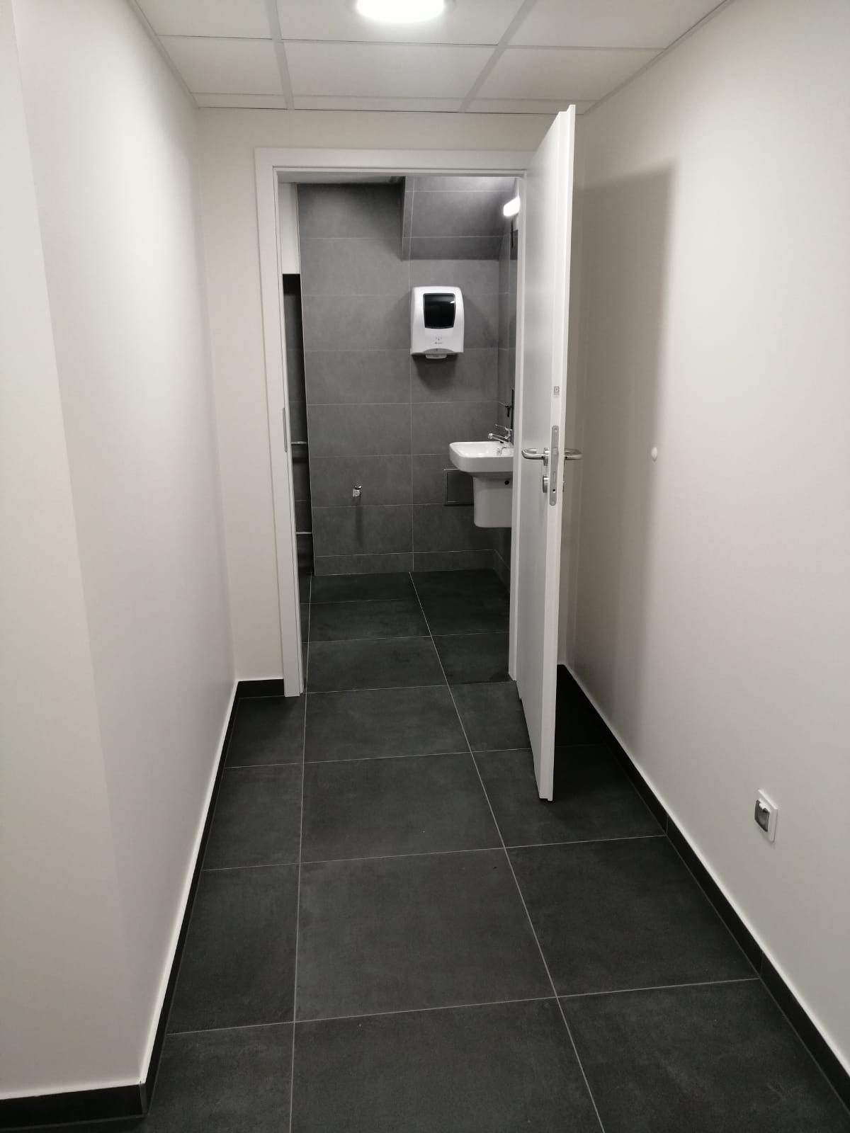 Nowoczesna łazienka z ciemnymi płytkami podłogowymi i szarymi płytkami ściennymi, biała umywalka i dozownik mydła, widok z korytarza przez uchylone białe drzwi.