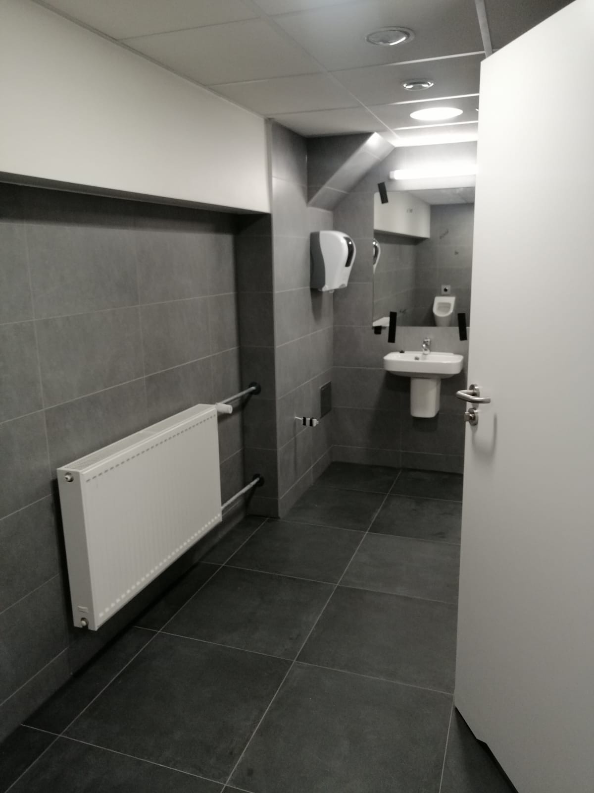 Szara łazienka w płytkach z grzejnikiem, umywalką i dozownikiem mydła, widok zza otwartych drzwi.