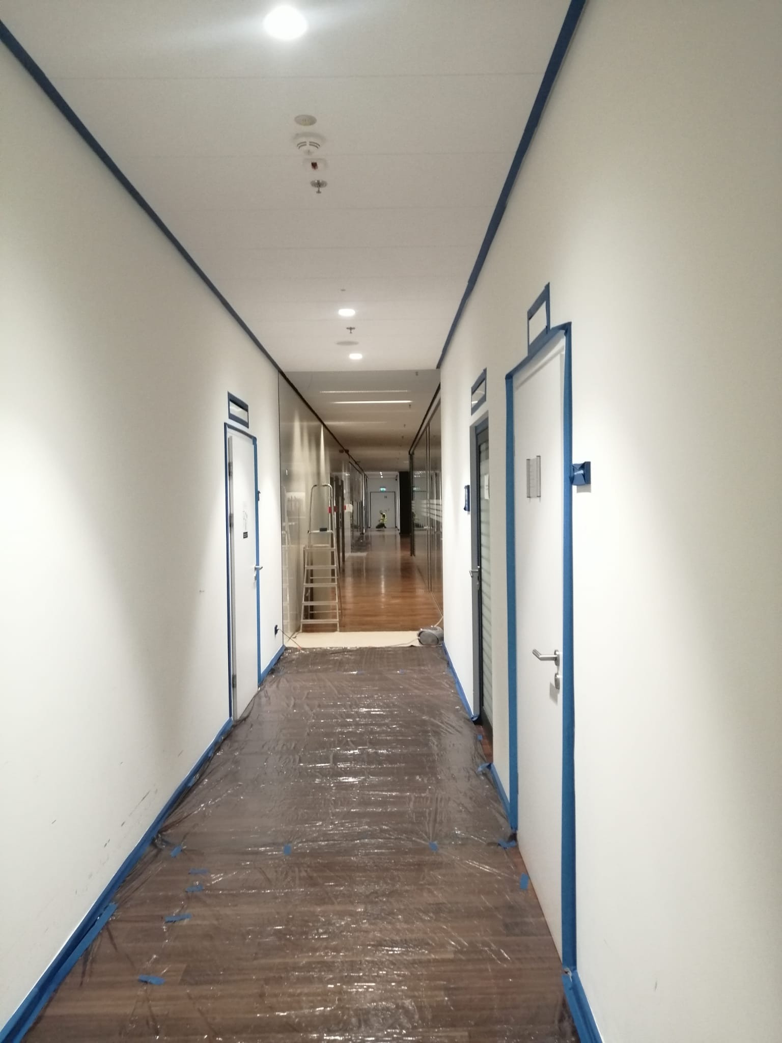 Korytarz biurowy przygotowany do malowania: ściany i drzwi oklejone taśmą malarską, podłoga zabezpieczona folią ochronną, drabina w tle.