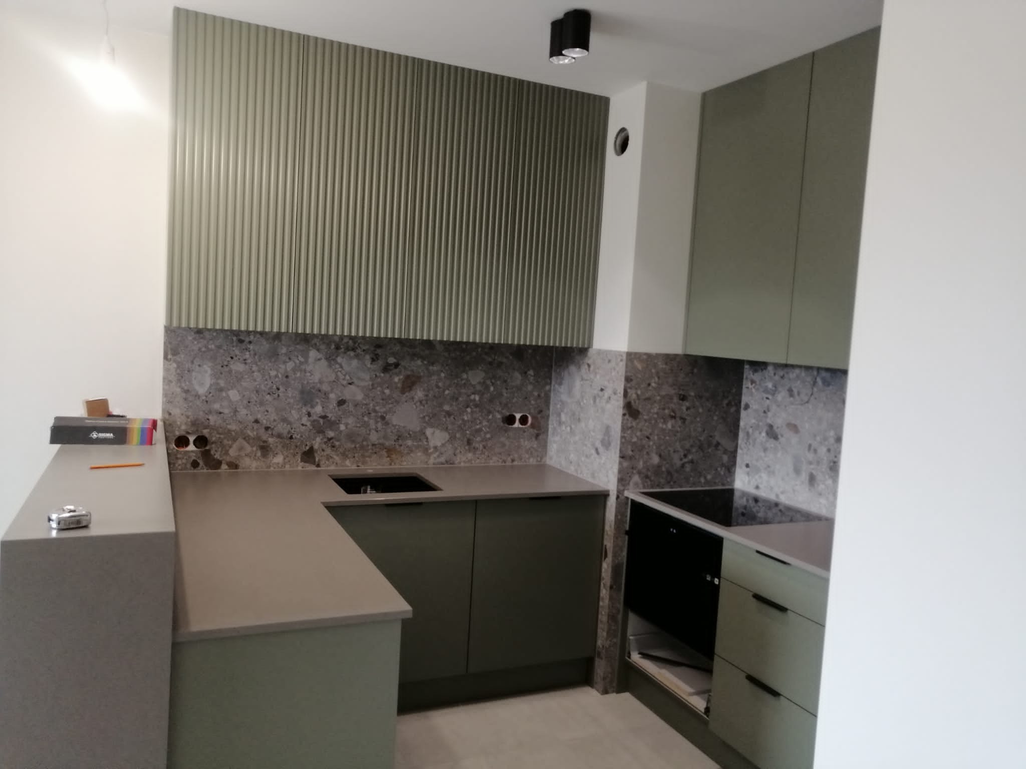 Nowoczesna kuchnia w odcieniach szarości i oliwkowej zieleni z ryflowanym panelem ściennym, kamiennym blatem i czarną płytą grzewczą.