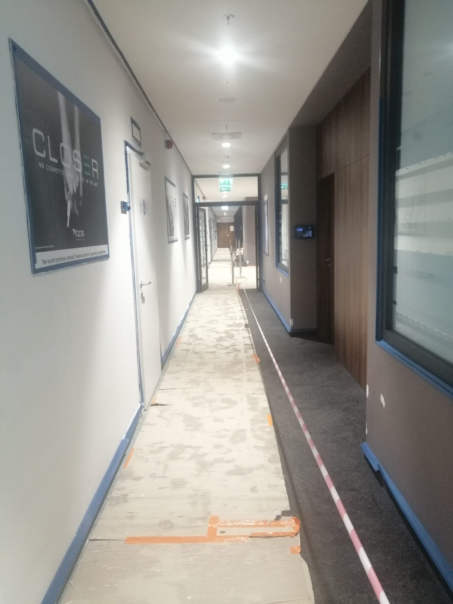 Korytarz biurowy w trakcie prac przygotowawczych do malowania: podłoga zabezpieczona kartonem, ściany oklejone taśmą malarską, widoczny plakat na ścianie.
